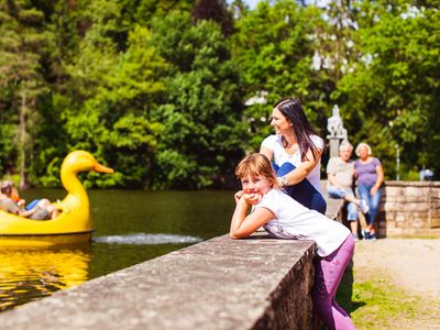 Das Bild zeigt ein kleines Mädchen im Vordergrund, das an einer Mauer gelehnt ist. Im Hintergrund guckt eine Frau aufs Wasser, auf dem ein Boot in Form einer gelben Ente fährt.