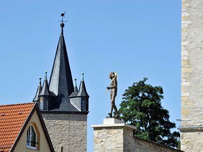 Das Bild zeigt einen Teil der Stadtbefestigung Ascherslebens mit Kirchturm, Statue auf einem Sockel und historischer Steinwand. 