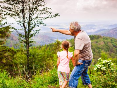 Das Bild zeigt ein kleines Mädchen mit seinem Opa, der ins Tal zeigt. Beide stehen an einer Aussicht im Sommer. 