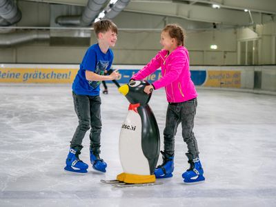 Das Bild zeigt einen kleinen Jungen und ein kleines Mädchen auf Schlittschuhen in einer Eishalle. Sie halten sich an einer Pinguinfigur fest. 